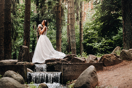 一位身着白色连衣裙 戴着手套 手捧花束的优雅新娘站在森林的溪流旁 享受着大自然 自然公园里穿着婚纱和手套的模特 白俄罗斯金发女郎图片