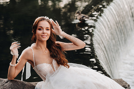 一位穿着白色连衣裙 手套和赤脚的优雅新娘坐在公园的瀑布附近享受大自然 自然公园里穿着婚纱和手套的模特 白俄罗斯婚姻婚礼若虫裙子女图片
