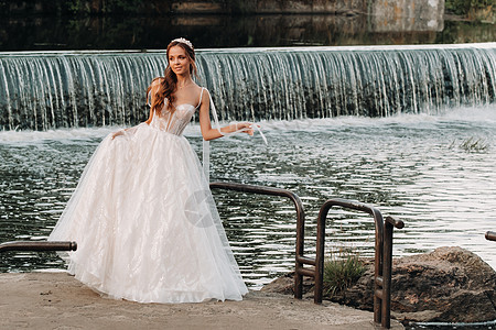 一位穿着白色连衣裙和手套的优雅新娘站在公园的河边 享受大自然 自然公园里穿着婚纱和手套的模特 白俄罗斯婚姻瀑布旅行男人女孩幸福若图片