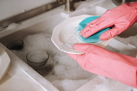 粉红色的盘子戴粉红色手套的家庭主妇女孩用洗涤剂在水槽里用手洗碗 女孩在家里打扫房子 戴着手套洗碗盘子海绵眼镜家务台面洗涤菜肴女士公寓卫生背景