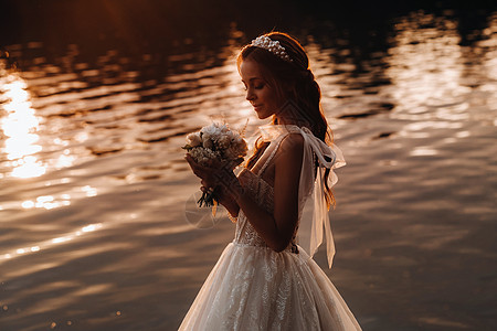 一位穿着白色连衣裙和手套的优雅新娘拿着花束站在公园的河边 在日落时享受大自然 自然公园里穿着婚纱和手套的模特 白俄罗斯幸福婚礼若图片