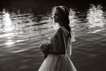 一位穿着白色连衣裙和手套的优雅新娘拿着花束站在公园的河边 在日落时享受大自然 自然公园里穿着婚纱和手套的模特 白俄罗斯 黑白照片图片