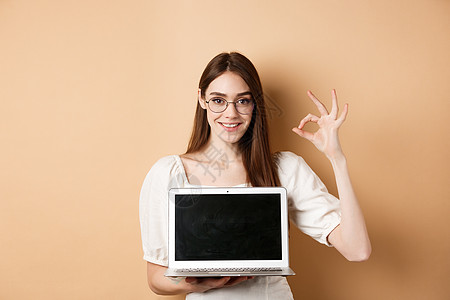 电子商务 戴着眼镜的微笑的年轻女性显示出好的标志和笔记本电脑屏幕推荐米色背景上的互联网宣传化妆品工作室褐色购物周年自由职业者女孩图片
