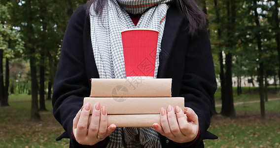 在秋季公园的背景下 一位身穿黑色外套和灰色围巾的年轻女子手里拿着一叠书和一杯咖啡 在露天 教育 女孩的近手叶子诗歌商业毛衣女士闲图片