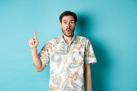 穿着夏威夷衬衫的caucasian旅游者惊讶得要命 举起手指说哇 看升职情况 站在蓝背景上站立工作室成人男性广告快乐生活假期胡子背景图片