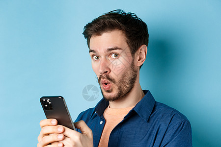 惊讶的白人男子的特写说哇 看着相机很开心 拿着智能手机 站在蓝色背景上工具促销成功电话工作室广告商业技术成人发型背景图片