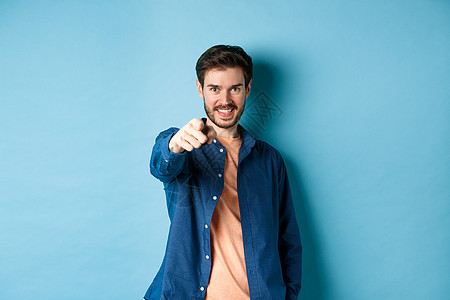自信的微笑的家伙选择或邀请你 用手指指着相机决定 站在蓝背景上站立生活快乐情绪广告胡子男人成人成功工作室胡须背景图片