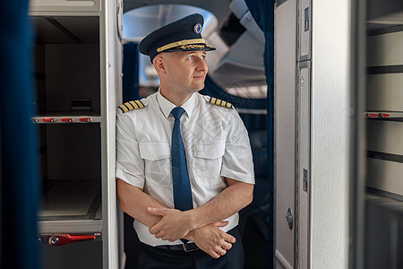 穿着制服和帽子的体贴男飞行员站在飞机内时双臂交叉并望向一边图片