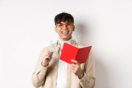戴着眼镜的快乐年轻人笑着记笔记 在计划表中写下 拿着笔和日记 站在白色背景上潮人黑发发型日程男人写作规划师学生工作室杂志图片