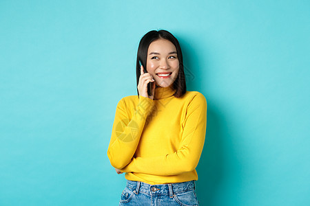 穿着黄色毛衣的有吸引力的韩国妇女 聊天和微笑 在手机上交谈 看左侧复制空间 站在蓝底边站立黑发情感广告商业学生快乐青少年女性手势图片