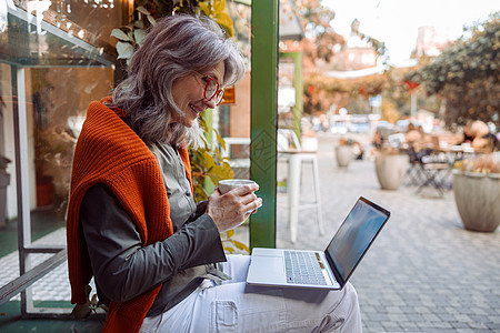 美丽的高龄妇女坐在咖啡馆附近的长椅上 在笔记本电脑上使用视频聊天机背景图片