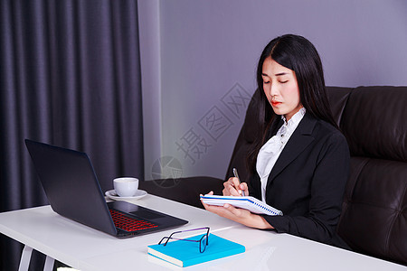 坐在桌上用笔记本电脑在笔记本上写字条的女商务人士图片