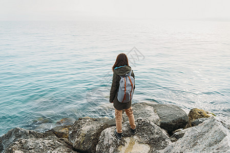 妇女站在海岸线的石头上背包海滩蓝色冒险女孩享受女性旅行游客远足者图片