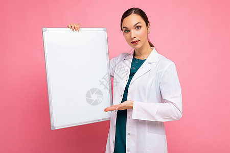 身穿医用白大衣 手持白色磁板的迷人漂亮年轻正面黑发女性肖像 在粉红色背景中进行模拟 并带有复制空间图片