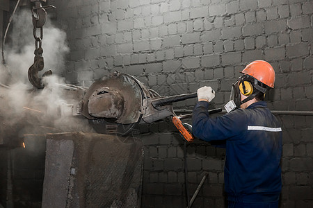 一名工人身着保护头盔 呼吸器和穿衬服 在工厂车间用重型设备投掷铁混凝土浴管碾磨;图片