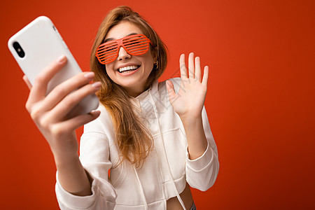 迷人迷人的年轻微笑快乐的女人手持和使用手机自拍 穿着时髦的衣服 与墙壁背景隔离眼镜社交互联网神器博主电话金发女郎白色摄影帽衫图片