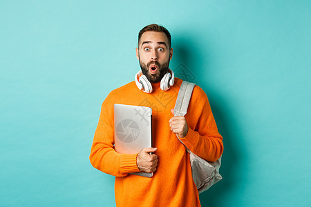背着背包和耳机的快乐男人 拿着笔记本电脑 微笑着 看起来很惊讶 站在绿松石背景上促销胡子橙子冒充胡须黑发横幅学生毛衣工作背景图片