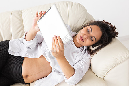 拉丁美洲怀孕妇女在家里躺在沙发上时使用平板电脑 怀孕和为人父母的概念信息等情况中药片工具阅读拉丁孩子生活肚子技术腹部母亲图片