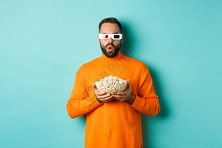 穿着橙色毛衣和3D眼镜的帅哥 看电影欢快 拿着爆米花 看着惊讶 站在蓝背景上促销标识黑发胡子男性横幅潮人广告情感快乐图片