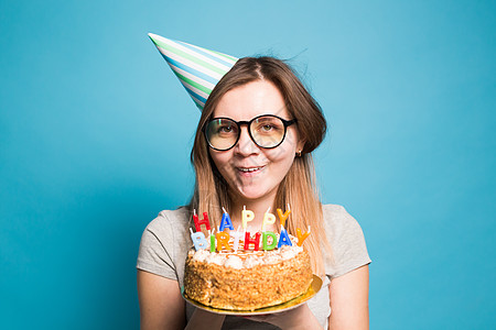 穿着纸帽和眼镜的疯狂可笑女孩 在蓝背景上拿着一个生日蛋糕 恶作剧和问候的概念 笑声惊喜节日蛋糕女士喜悦生日甜点派对故事情绪图片