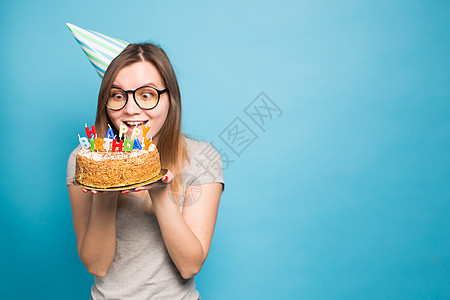 穿着纸帽和眼镜的疯狂笑女 在蓝色背景上拿着一个大生日蛋糕 带复制空间 恶作剧和问候的概念图片