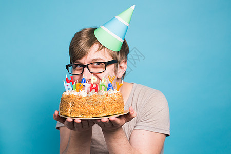 戴着问候纸帽的滑稽疯狂年轻人想咬掉一块祝贺蛋糕 鬼混和生日祝福的概念 复制空间庆典周年工作室派对节日蓝色海报礼物创造力乐趣图片