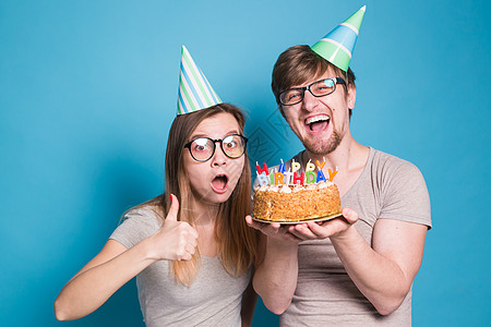 可爱的年轻情侣迷人帅哥 和穿着纸帽的可爱女孩 做傻脸 手里拿着一个生日蛋糕 站在蓝色背景上周年风格蛋糕情绪惊喜男人甜点礼物生日创图片
