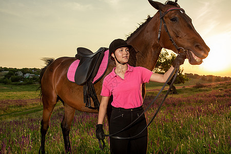 美丽的微笑女孩骑师站在她棕色的马旁边 在天空和绿色田野背景上穿着特殊制服在日落女性马背马术动物抑制农村女孩外套运动活动图片