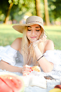 年轻的caucasian年轻女人 戴帽子保持羊角面包 躺在公园 在格子和阅读书上图片