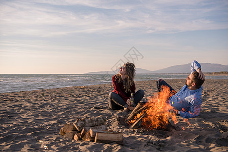 春火旁的海滩上坐着年轻夫妇 喝啤酒篝火女性靴子友谊啤酒喜悦男生男性瓶子火焰背景图片