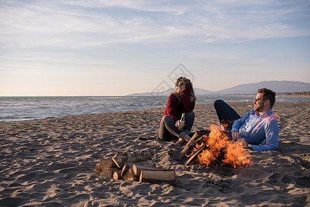 春火旁的海滩上坐着年轻夫妇 喝啤酒友谊喜悦夫妻海洋烹饪假期瓶子男人闲暇女性背景图片