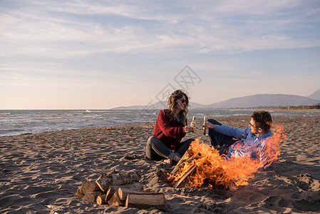 春火旁的海滩上坐着年轻夫妇 喝啤酒闲暇毛衣瓶子夫妻烹饪女性海洋旅行火焰篝火图片