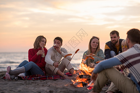 坐在海滩火炉边的年轻朋友团体香肠烧烤旅游火焰烹饪电话饮料野餐乐趣青年图片