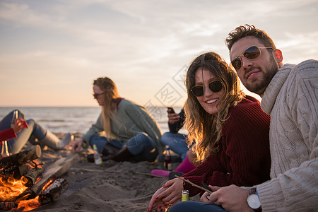 在海滩日落时和朋友共度欢乐的情侣啤酒营火休闲夫妻火焰沙滩乐趣衣服快乐动作图片