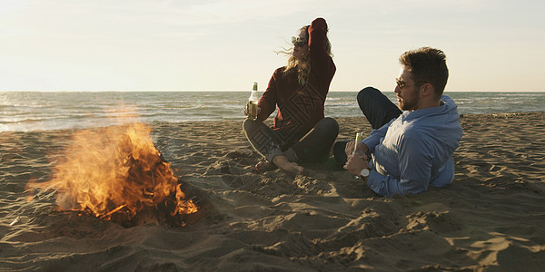 爱的年轻情侣坐在露营火边海滩上喝啤酒男性女性男人烹饪喜悦男生海洋闲暇篝火女朋友图片