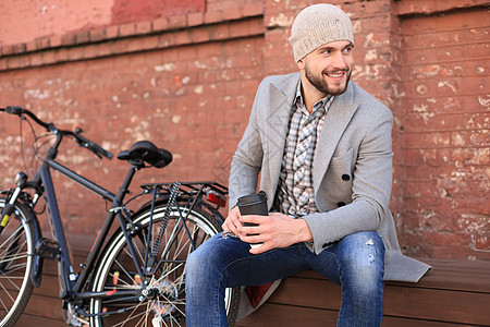 穿着灰色大衣和帽子的帅哥坐在长椅上 喝着咖啡 想着自己的自行车思维运动太阳运输成人牛仔裤太阳镜金发女郎拉丁城市图片