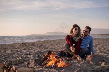 春火旁的海滩上坐着年轻夫妇 喝啤酒靴子女性友谊夫妻火焰毛衣海洋喜悦啤酒男性背景图片