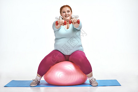 红发 胖胖女人坐在球上 保持哑铃声女性锻炼乐趣重量腹部哑铃运动健身房身体肥胖背景