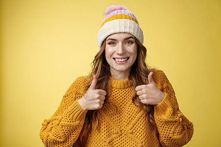 特写迷人的支持善良的年轻女性竖起大拇指 微笑着赞同你的想法 鼓励跟上 满意的好选择 站着高兴的黄色背景快乐学生温暖大学新年帽子名图片