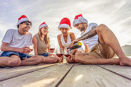 快乐的人在异国情调的度假胜地喝酒享受户外生活 一群年轻朋友在日落时一起玩乐 戴着红色圣诞帽坐在海上度假胜地的码头上图片