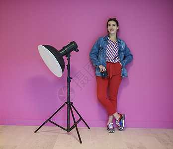 年轻女士一边靠着粉红色墙 一边拿着照相机倾斜成人技术手电筒相机职业工具女孩冒充工作图片