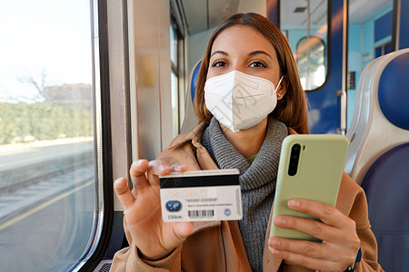 充满自信的有FFFP2 KN95保护面罩的妇女 带着火车旅行 观看持有信用卡和智能电话的照相机图片