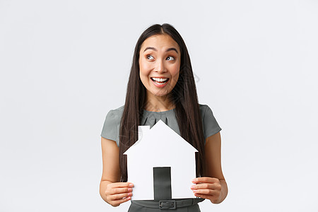 保险 贷款 房地产和家庭概念 微笑快乐的亚洲女人找到了完美的家 拿着纸房子 欢欣鼓舞 看着横幅的左上角 白色背景 看起来很有趣企图片