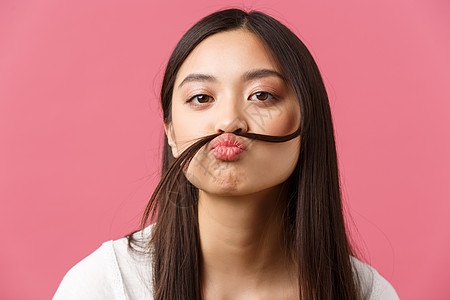美丽 人们的情感和夏季休闲概念 有趣而无聊的愚蠢亚洲女孩的特写 她在嘴唇上留着假胡子 盯着镜头嬉戏 粉红色背景图片