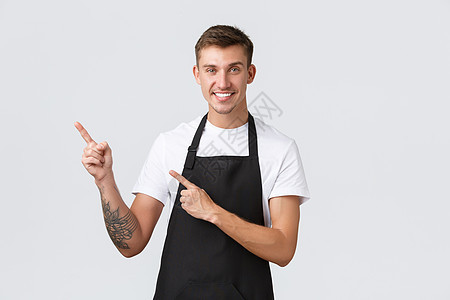 小企业主 咖啡店和员工的概念 帅气友善的咖啡师 身穿黑色围裙的男人向左指指点点 微笑着邀请在咖啡厅享用饮品 白色背景图片