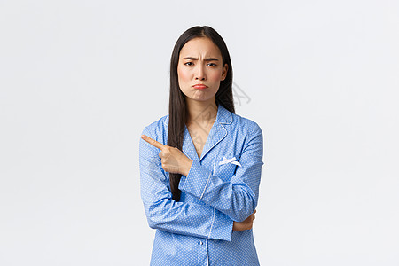 穿着蓝色睡衣的沮丧阴郁的亚洲女孩失望而悲伤地做着鬼脸 表现出一些不好的东西 向左指指点点 噘嘴 抱怨不公平或悲伤的事情 站在白色图片