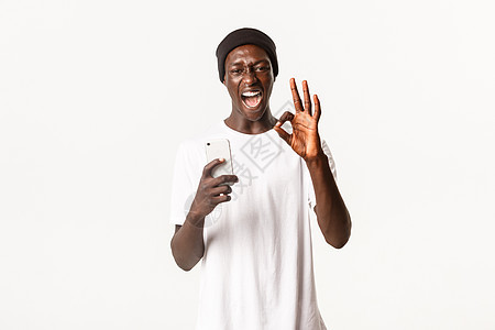 使用手机 满足满意 站立白背景 展现出像应用程序一样的好手势的非洲-美籍年轻人的平凡肖像图片