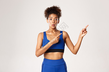 锻炼和健身的概念 穿着蓝运动服 在右上角指手指 白背景的非洲-美国女运动员感到惊讶和好奇 并举足轻重图片