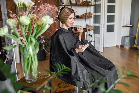 快乐的金发碧眼美女坐在椅子上 在理发厅等美容师时 正在使用智能手机女性客户镜子成人沙龙女孩男人工作拉丁剪刀图片