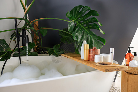 用于美容和身体护理产品的木架 位于现代白色泡泡浴缸上方的木架上 用绿色热带植物龟背竹装饰的舒适时尚的浴室瓶子肥皂淋浴洗发水凝胶架图片